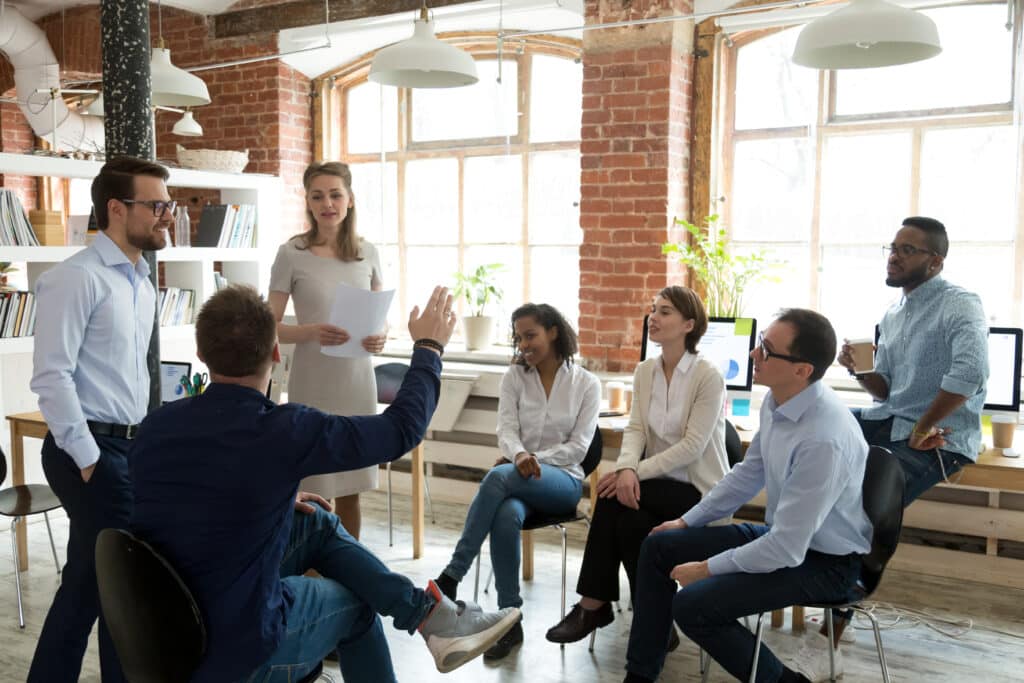 Un groupe de 3 femmes et 4 hommes formant un cercle assistent à une formation au leadership dans une grande salle moderne en briques rouges et l'un des hommes de dos lève la main pour prendre la parole.