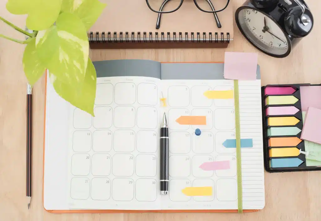 Un agenda est ouvert sur une table en bois avec un stylo dessus, des post-it colorés, un réveil, un crayon, un carnet, une plante verte et des lunettes à côté.