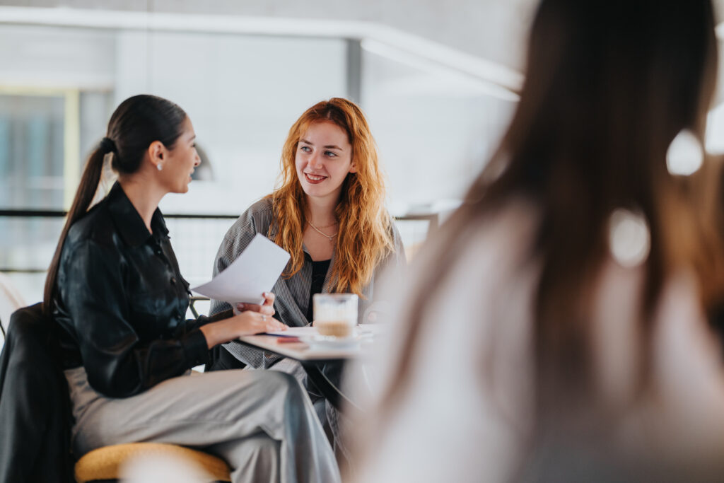 relaxed business conversation between two women at a coffee shop, with documents and coffee