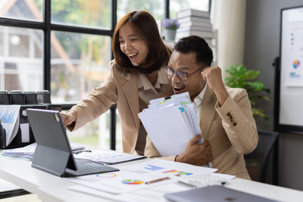 surprised businesspeople celebrating project success while looking at a tablet, sharing excitement over positive financial report results and corporate achievements.