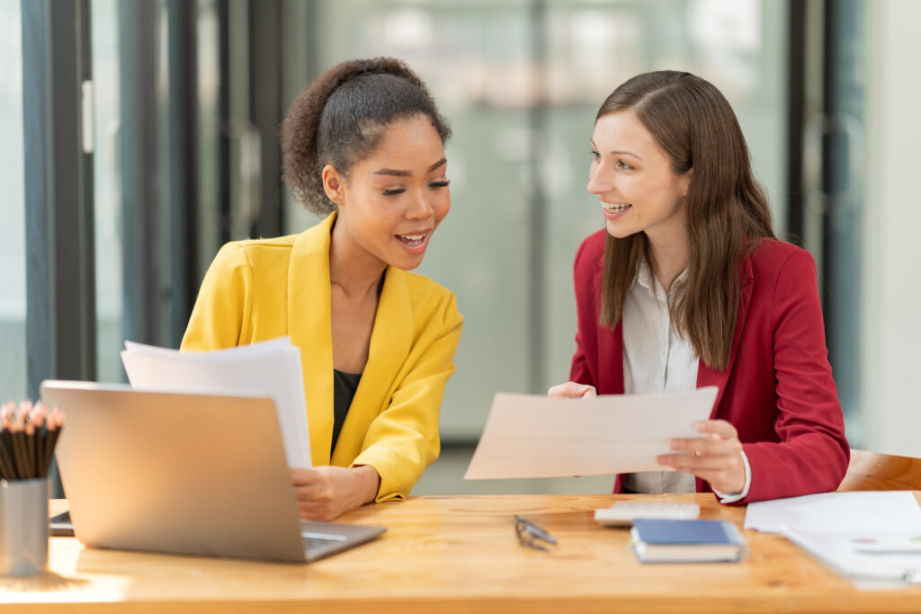 two smiling multi ethnic businesswomen reviewing documents and discussing data at a wooden desk with a laptop, highlighting teamwork and professional collaboration in a contemporary workspace